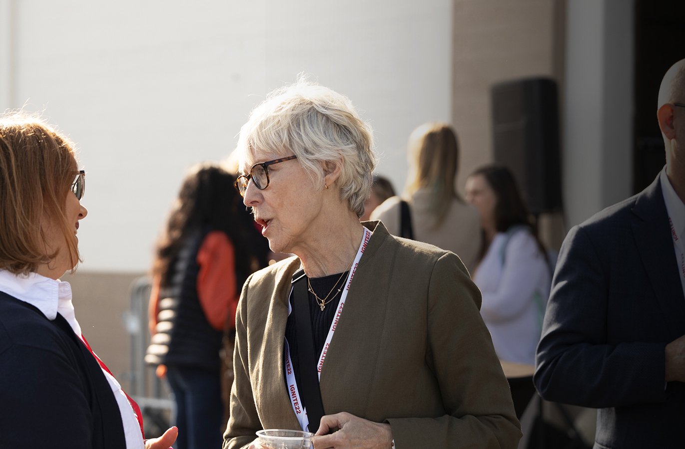 A young black woman and a white man speak in the foreground, with a diverse audience gathered in the background at an event.